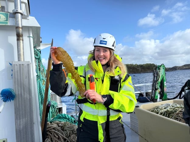 Kvinne i gult sikkerhetsutstyr med hjelm på. Hun holder opp en tarebit og smiler til kamera. 