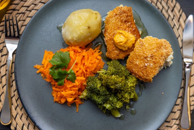 Sett ovenfra: et blått fat med raspet gulrot med grønt bladkrydder på, en kokt potet, to stykker fiskegrateng, hvor den ene har en dæsj oransje kryddersmør oppå og noen kokte brokkoli. 